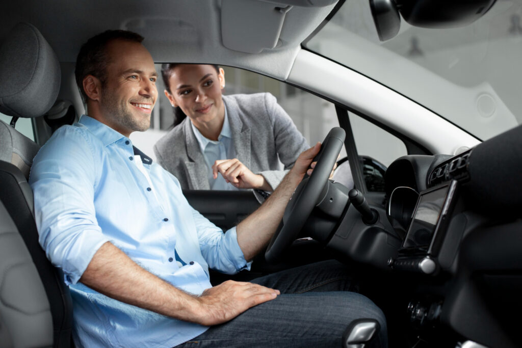 A friendly female private driving instructor standing outside the car door, guiding a smiling adult student during their first hands-on driving lesson for beginners.