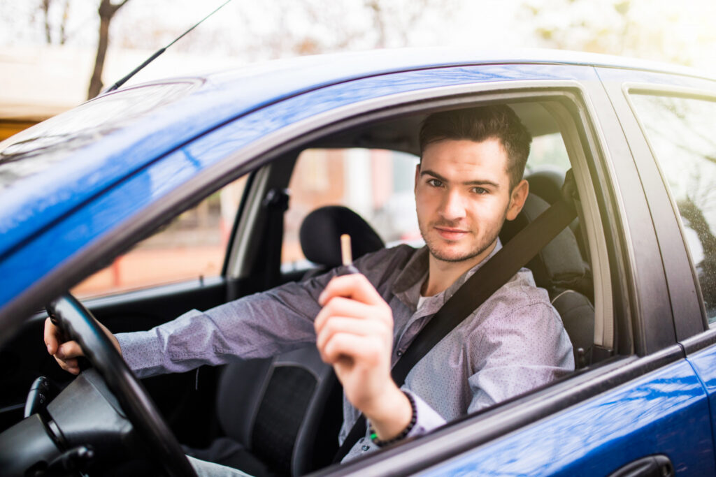 A happy young man sitting behind the steering wheel of a blue car holding up a car key, celebrating passing his G2 driving test.