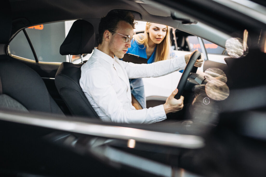 A male student with glasses practice steering controls while a female driving instructor looks inside the car window during in-car driving lessons.