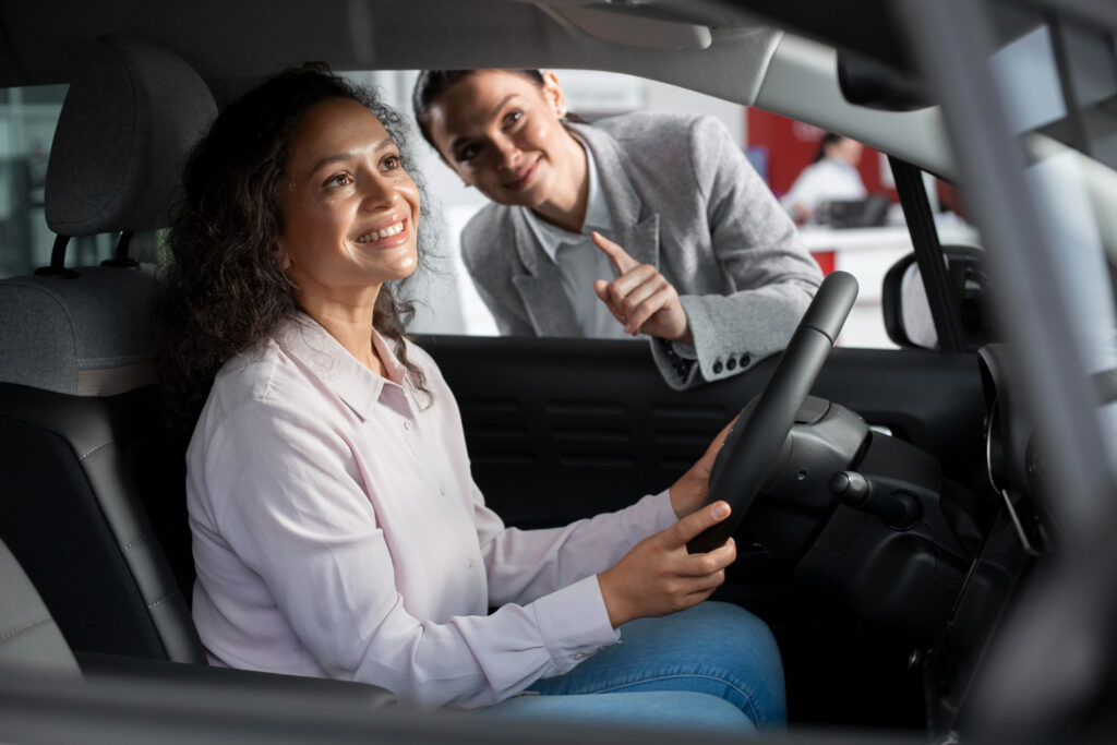 A friendly female private driving instructor standing outside the car door, guiding a smiling adult student behind the steering wheel during an in-car driving lesson.