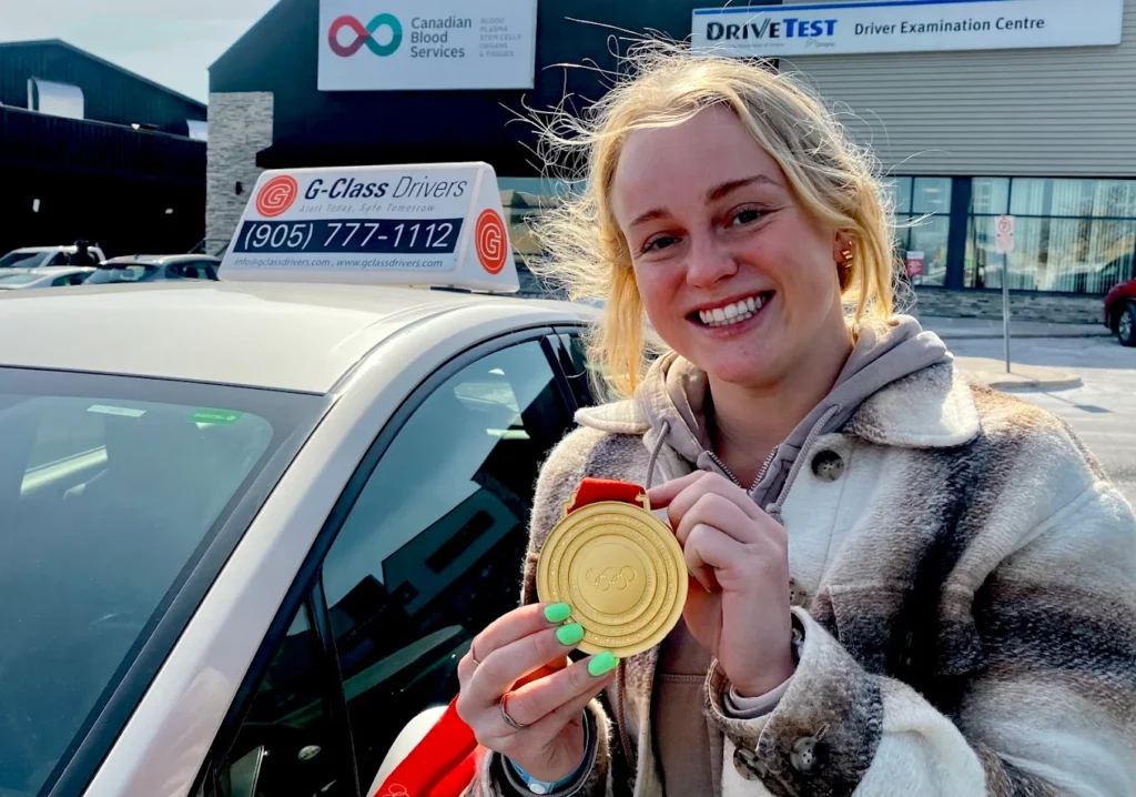 A smiling young female Olympic gold medalist holding up her medal next to a G-Class Drivers training car at an Ontario DriveTest Centre.