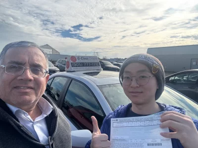 A driving instructor standing next to a happy student holding their official Ontario temporary driver's license paper after passing the road test with a G-Class Drivers training car.